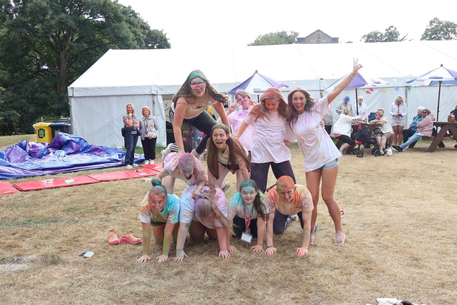 A group of young people in a human pyramid on the grass, dusted with coloured powder, with a marquee behind them