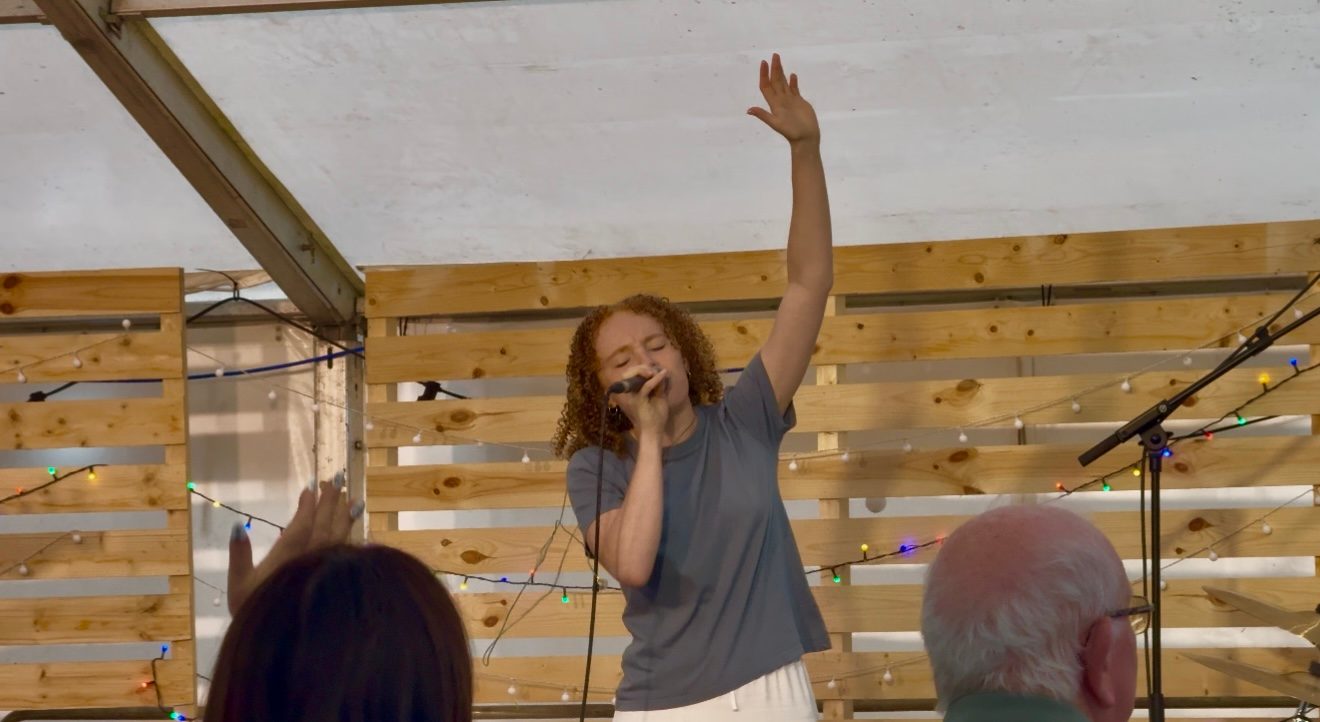 A worship leader singing with one hand raised under a wooden shelter, with people listening in the foreground